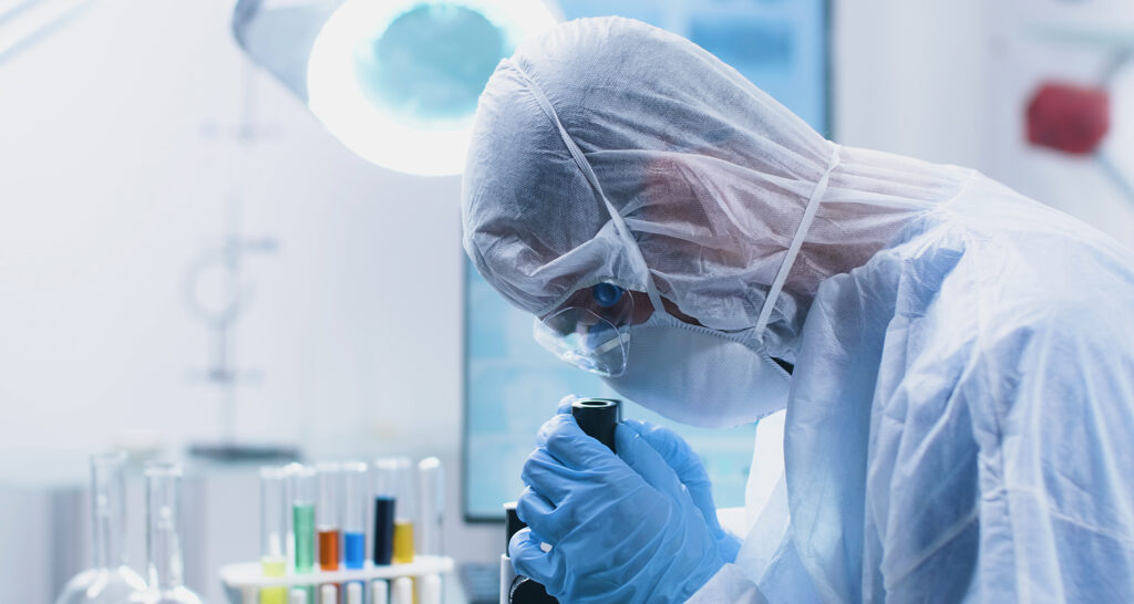 Researcher doctor with ppe equipment looking into medical microscope analyzing virus sample during vaccine development experiment in biochemistry hospital laboratory. Medicine concept